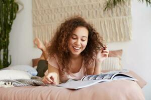 Portrait of young laughing dark skinned woman with curly hair, lies on the bed and reads a cool newspaper article at magazine,broadly smiles and enjoy sunny free day. photo
