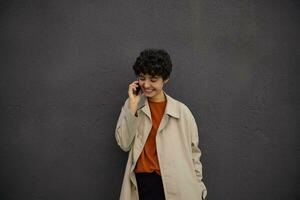 al aire libre Disparo de joven Rizado morena mujer con corto Corte de pelo en de moda atuendo haciendo llamada con su móvil teléfono mientras caminando terminado ciudad calles, posando terminado negro urbano pared en elegante ropa foto