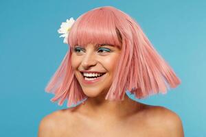 Studio shot of cheerful young beautiful woman wearing white flower in her short pink hair while posing over blue background, laughing happily while looking aside photo