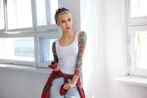 Indoor shot of young lovely tattooed blonde lady dressed in casual clothes while standing in front of big window on bright sunny day and looking attentively at camera photo