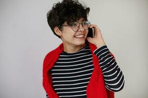 Cheerful young pretty dark haired curly woman with short trendy haircut dressed in stylish casual wear while posing over white background, smiling sincerely with mobile phone in raised hand photo