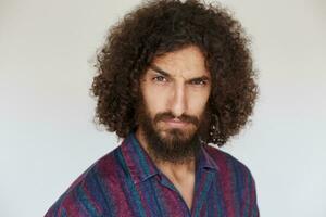 Portrait of stern brunette bearded guy with dark curly hair raising eyebrow and looking severely at camera, keeping lips folded while posing against white background in casual shirt photo