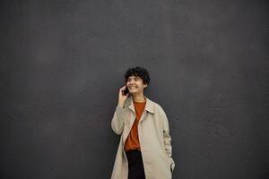 Happy beautiful young dark haired female with short haircut keeping smartphone in her hand and having pleasant talk, having nice mood and smiling happily while posing over city environment photo