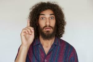 Studio shot of excited handsome young dark haired curly male with beard raising forefinger as he is having good idea, wrinkling forehead with wide eyes opened against white background photo