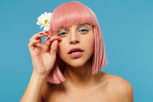 Close-up of young lovely pink haired lady with bob haircut adjusting her festive makeup with raised hand while posing over blue background with white flower in her hair photo