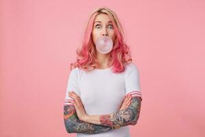 Young surprised beautiful pink haired woman with tattooed hands, wears in white t-shirt, blows gum ball, looking to the left in surprise, stands over pink background. photo