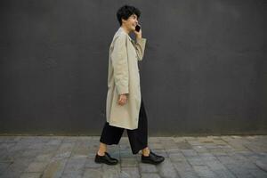 Side view of fashionable young pretty brunette woman walking on city street in stylish clothes, having nice talk on phone with her friend, being in high spirit photo