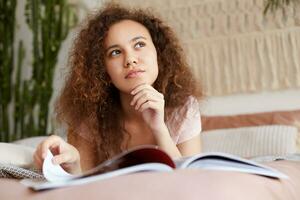 Portrait if dreaming young rested african american girl with curly hair, lies on the bed and reads a new issue of magazine, calmed looks away and touches chin. photo