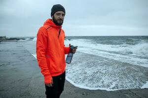 Outdoor photo of young brunette bearded runner with fitness bottle in raised hand looking aside and smiling slightly while posing on stormy weather over beach in warm athletic clothes