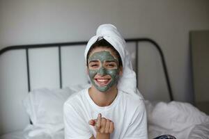 Happy young pretty brunette woman with mask on her face wearing white towel on head while sitting on bed over home interior, looking happily to camera and smiling broadly photo