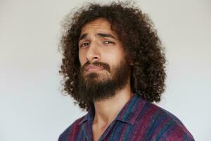 Portrait of handsome young bearded man with dark curly hair frowning eyebrows and wrinkling forehead while looking at camera, posing against white background in casual striped shirt photo