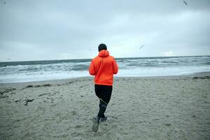 Back view of fitness male model working out and running by seaside on gray early morning, dressed in warm sporty clothes and orange coat. Sport and healthy lifestyle concept photo