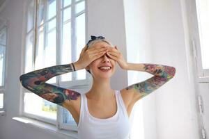 Good looking young lovely glad blonde woman with tattoos keeping raised palms on her eyes and smiling happily while standing over bright studio in white shirt photo