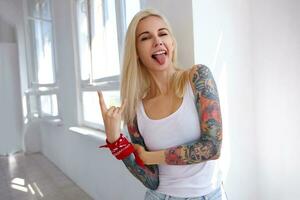 Funny shot of young tattooed pretty blonde lady with natural makeup showing rock and roll gesture and sticking cheerfully out her tongue, posing over bright studio photo