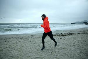 Shot of young bearded athletic male dressed in warm sporty clothes running on coastline of sea in early gray morning, looking ahead with concentrated face and keeping hands at chest level photo