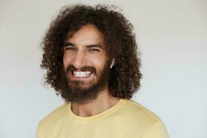 Happy attractive young brunette curly man showing his pleasant emotions while posing against white background, demonstrating his white perfect teeth while smiling broadly photo