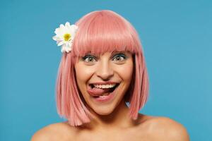 Funny shot of joyful young attractive female with short pink hair sticking out her tongue while looking cheerfully at camera, wearing colored makeup while posing over blue background photo