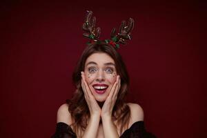 Surprised blue-eyed young brown haired woman with evening makeup wearing holiday hoop and looking at camera with wide eyes opened, keeping palms on cheeks and smiling widely photo