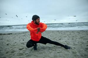 Outdoor shot of attractive active young bearded male keeping hands raised in front of himself while stretching his muscles before running exercises, isolated over seaside view photo