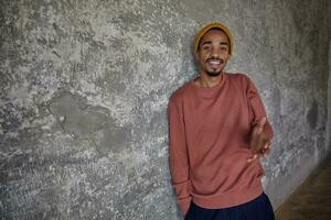 Positive pretty bearded dark skinned guy dressed in pink sweater and mustard cap looking to camera with light smile and raising palm up, posing against concrete background with hand in pocket photo