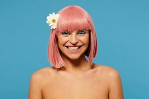 Happy young beautiful pink haired woman with bob haircut looking cheerfully at camera with wide charming smile, standing over blue background with white flower in her hair photo