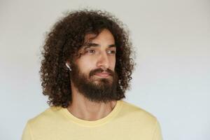 Handsome young bearded man with brown curly hair looking aside thoughtfully while listening to training with earphones, dressed in casual clothes while posing over white background photo