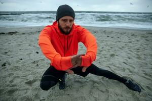 Image of young bearded male with eyebrow piercing looking at camera with serious face while stretching his muscules before running session, posing over seaside view photo