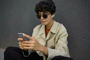 Close-up of attractive curly brunette woman with short haircut keeping mobile phone in hands and checking her mail box, keeping lips folded while sitting over black urban wall photo