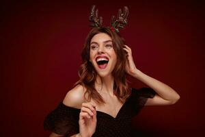 Indoor photo of beautiful joyful young brunette lady with festive hairdo wearing chrismas horns and elegant clothes while posing over claret background, expressing true positive emotions
