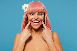 Studio photo of joyful young green-eyed woman with short pink haircut keeping forefingers on mouth corners while smiling happily at camera, isolated over blue background