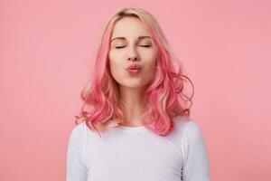 retrato de encantador positivo hembra con rosado Rizado pelo posando terminado rosado antecedentes y sonriente atentamente, acuerdo ojos cerrado y plegable labios en beso, vistiendo casual blanco camiseta foto