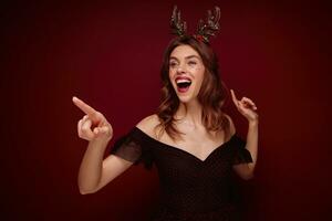 Christmasnew year themed portrait of young lovely brown haired woman in black elegant dress with red dots pointing cheerfully aside with forefinger and smiling widely, isolated over claret background photo