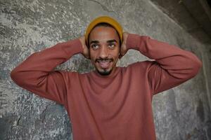 foto de positivo joven oscuro desollado hombre con marrón ojos cubierta su orejas con elevado palmas y smilling alegremente mientras mirando aparte, teniendo divertido con su amigos terminado hormigón antecedentes