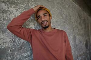 Puzzled young bearded dark skinned male with pretty brown eyes dressed in casual clothes, holding his head and frowning eyebrows while looking ahead, posing over concrete background photo