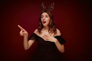 Open-eyed young beautiful brunette lady with festive hairstyle dressed in black girlish dress and christmas hoop showing aside amazedly with wide mouth opened, isolated over claret background photo
