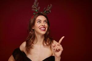 Happy young brown haired cheerful woman with festive makeup and silver stars on her face showing cheerfully upwards with forefinger, enjoying x-mas theme party over claret background photo