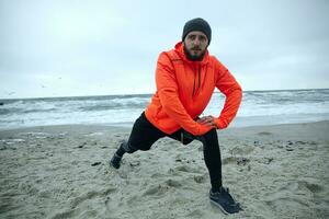 Horizontal shot of attractive young bearded male stretching his muscles before running exercise, standing over coastline of sea on gray cold weather, goes in for sport every morning photo