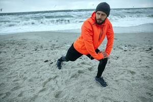 Pensive young brunette bearded runner stretching his muscles before running session, standing over seaside on gray stormy morning, starting day from morning jog photo