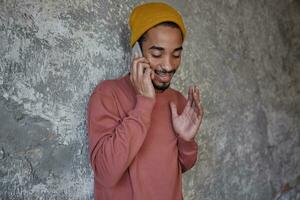 Indoor photo of positive pretty young dark skinned male with beard keeping mobile phone in hand and making call to his friend, standing over concrete wall with raised hand