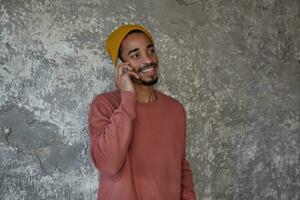 Cheerful bearded dark skinned male with charming brown eyes looking aside and smiling happily while having nice talk on phone, posing over concrete background in casual clothes photo