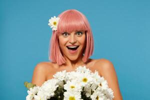 Indoor photo of surprised pretty young woman with short pink haircut looking excitedly at camera with wide mouth opened, receiving flowers from secret boyfriend, isolated over blue background