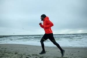 imagen de joven hermoso barbado deportista escuchando a favorito música lista mientras trabajando fuera y corriendo por playa en temprano nublado Mañana. aptitud y deporte concepto foto