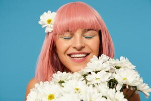 Good looking young happy woman with short pink hair wearing festive makeup while posing in white flowers over blue background, smiling pleasantly with closed eyes photo