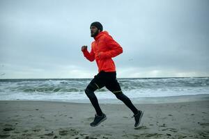 Side view of young sporty brunette bearded man in warm orange coat and black athletic clothes exercising outdoors before working day, goes in for sport every morning. Fitness and sport concept photo