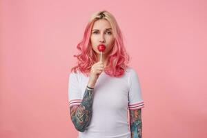 Indoor photo of pretty young tattooed woman with pink curly hair looking at camera with raised eyebrow, standing over pink background and keeping candy in her mouth