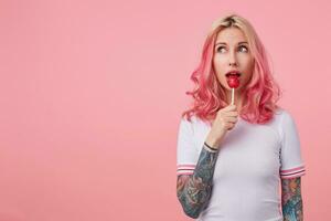 retrato de atractivo joven tatuado dama con rosado Rizado pelo mirando aparte soñando, posando terminado rosado antecedentes en casual blanco camiseta y paliza caramelo en palo foto