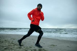 Photo of young bearded man in good physical shape looking over his shoulder while doing morning run at the seaside. Male fitness jogging training wellness concept