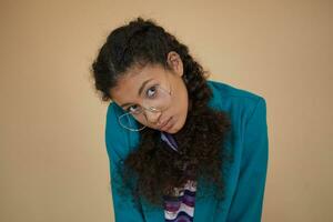 Shot of young curly brunette dark skinned lady with braids looking to camera with calm face and keeping lips folded, dressed in turquoise blazer and striped sweater while posing over beige background photo