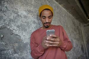 Close-up of handsome young bearded man with dark skin holding mobile phone in raised hands and looking on screen with concentrated face, posing over concrete wall in casual clothes photo