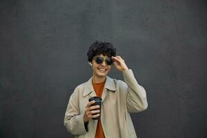 Positive smiling pretty young brunette woman with short curly hair standing over black concrete wall, holding her sunglasses and going to drink takeaway coffee, wearing stylish outfit photo